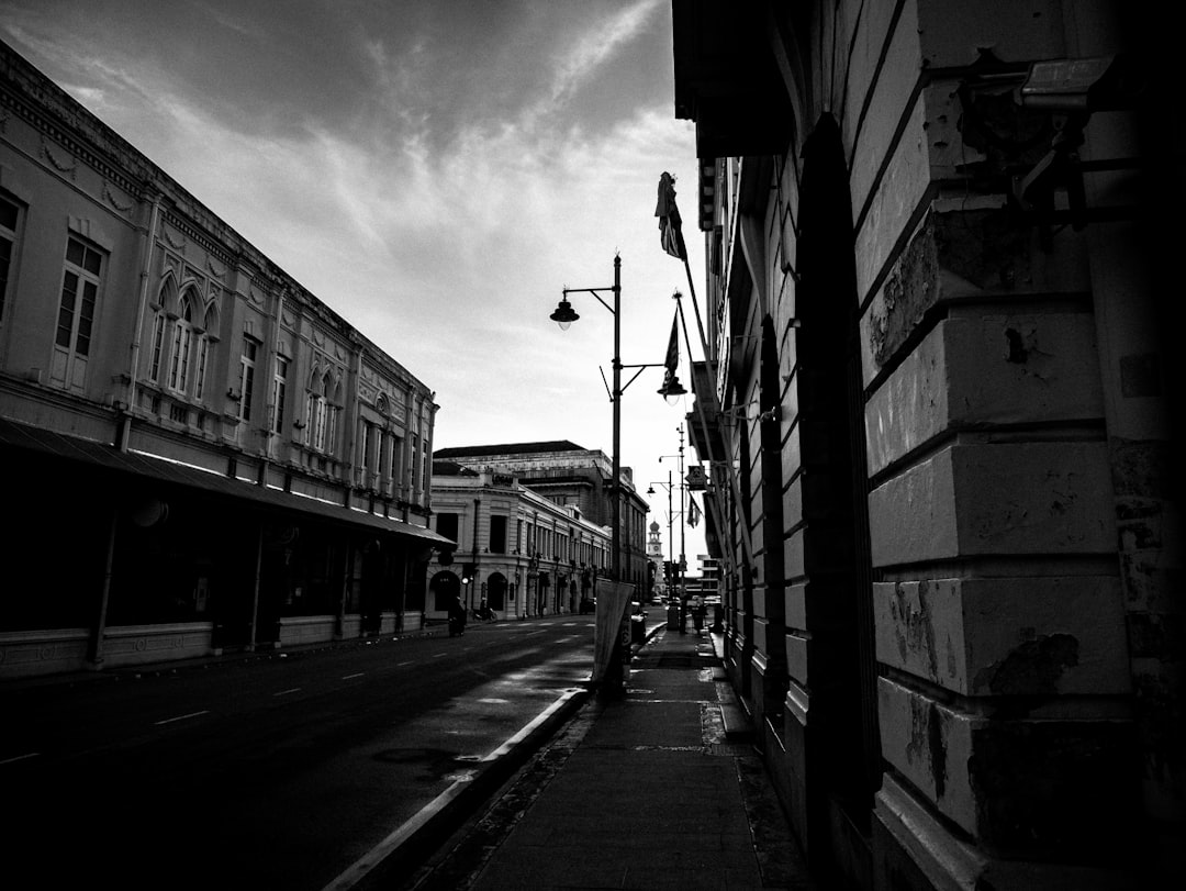 A black and white street photograph of the streets in “Bhive”, New Zealand, a place where hip hop meets colonial architecture. The city is filled with historic buildings that have been converted into art markets or fruit stands. A lone person walks down an empty sidewalk. The sky above is a dark grey, with light from the sun shining through clouds. The mood is somber yet hopeful. The feeling in your heart must be one of mystery and determination. The atmosphere is tense as you wonder what lies ahead, in the style of [Banksy](https://goo.gl/search?artist%20Banksy). –ar 4:3