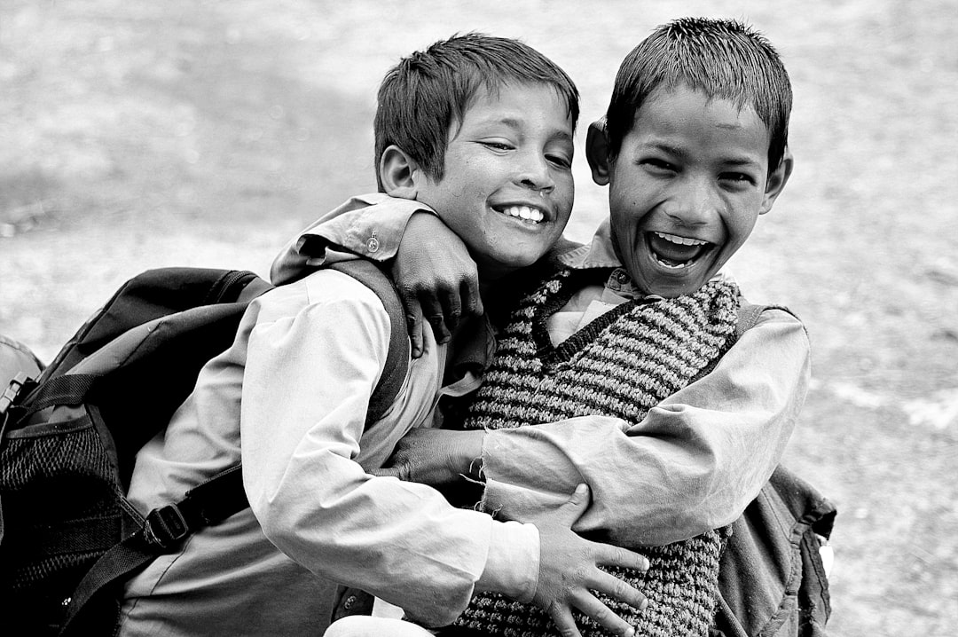 Black and white photo of two happy boys hugging. One is wearing an Indian dress with a vest over it, the other boy has on jeans, a shirt, and a backpack. The setting appears to be outdoors or in front of a school, as they appear from behind their backpacks. They both have black hair and bright eyes, laughing joyfully. Their expressions convey happiness and friendship. In between them there’s another person who seems slightly out of frame, adding depth to the scene. –ar 128:85