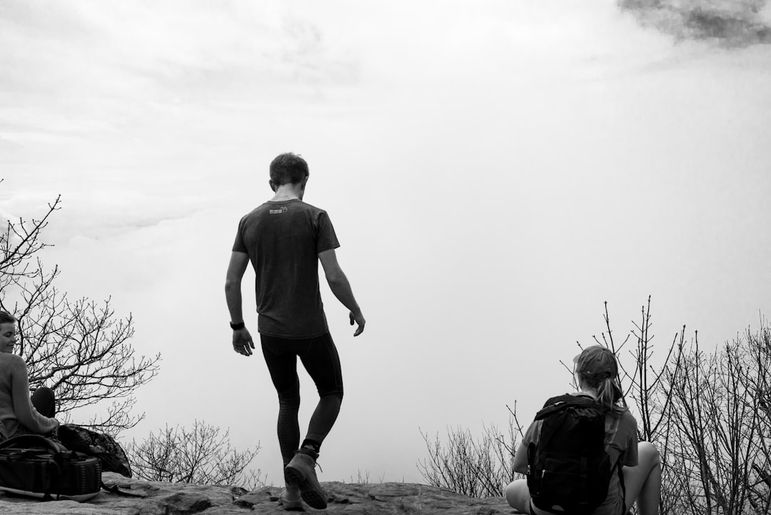 Black and white photography of a fit man in sportswear, hiking with friends on the Blue Ridge Mountain in West Virginia USA. The scene captures him standing at his best while enjoying nature’s beauty. He is wearing a t-shirt and black leggings. His relaxed posture reflects contentment as he gazes out over the horizon from atop the blue ridge mountain. In front of them, two women sit down resting after climbing up to their total height. It’s foggy but beautiful. –ar 128:85