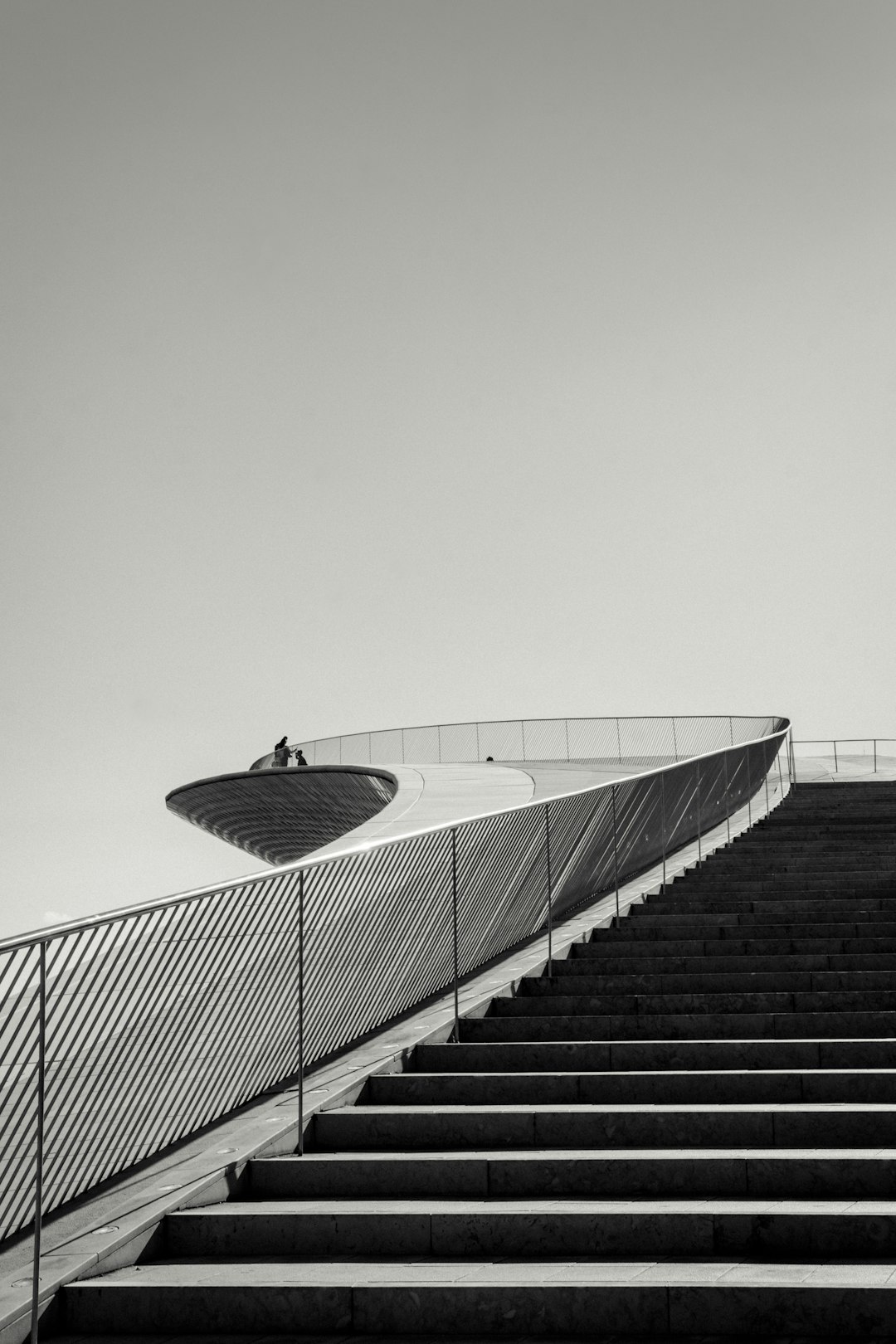 black and white photo of the minimalist futuristic architecture of a child’s park in Barcelona, with concrete stairs leading up to an oval shaped roof on top, with metal railings. There is one person sitting at the edge looking out over the city below. The sky above should be clear and bright, creating strong contrast between light grey and dark black tones in the style of [Ansel Adams](https://goo.gl/search?artist%20Ansel%20Adams). –ar 85:128