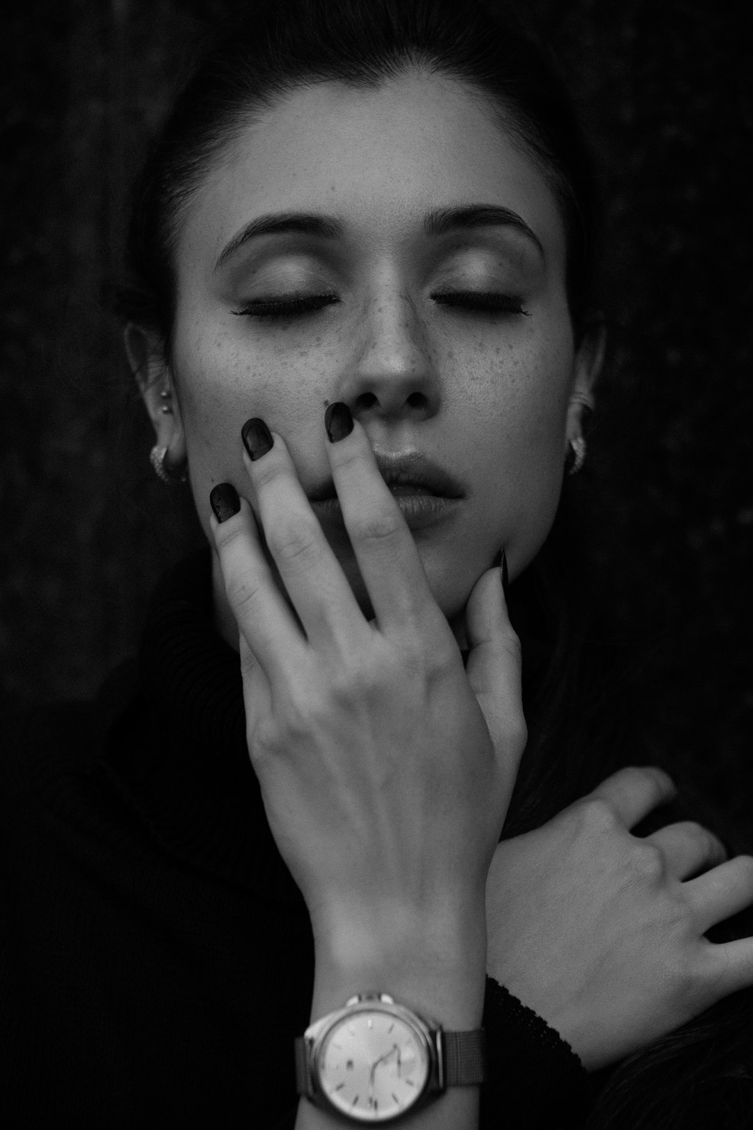 Black and white photography of an elegant woman with her eyes closed, wearing a watch on one hand while holding another hand near the face. Her nails have black nail polish. The photo is taken from below. –ar 85:128