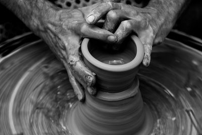 A black and white photograph of hands crafting an intricate clay vessel, capturing the motion blur as they spin on their potter's wheel. The focus is sharp with details in each curve and line of the masterpiece. This shot emphasizes a fine art photography style, highlighting every texture of clay against stark contrast to highlight its craftsmanship. --ar 128:85