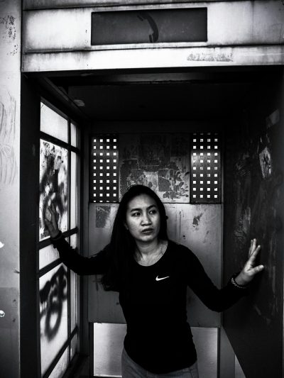 An Indonesian woman in her thirties, wearing a black Nike t-shirt and jeans, is standing inside an open phone booth with one hand outstretched as if to make the call she is about to place. The photo was taken from behind so we can see that there is graffiti on the walls of the phone booth. Her expression looks worried or nervous. The lighting gives it a cinematic quality and it is black and white photography. Shot in the style of Arriflex camera using Canon K35 prime lenses. --ar 3:4