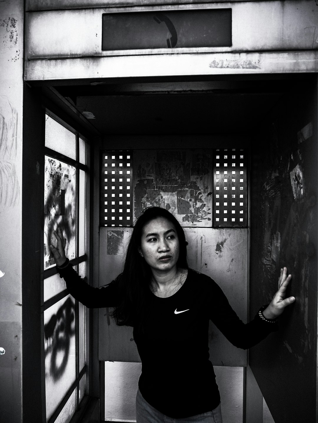 An Indonesian woman in her thirties, wearing a black Nike t-shirt and jeans, is standing inside an open phone booth with one hand outstretched as if to make the call she is about to place. The photo was taken from behind so we can see that there is graffiti on the walls of the phone booth. Her expression looks worried or nervous. The lighting gives it a cinematic quality and it is black and white photography. Shot in the style of Arriflex camera using Canon K35 prime lenses. –ar 3:4