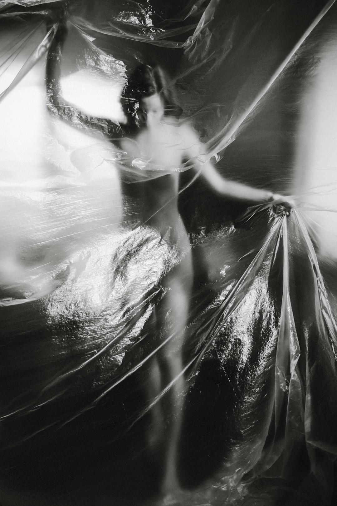 Abstract blurry black and white photograph of a female dancer wrapped in plastic sheeting, appearing ethereal. The photograph is in the style of an abstract blurry black and white image in the manner of plastic sheeting wrapping a female dancer, with an ethereal quality. –ar 85:128