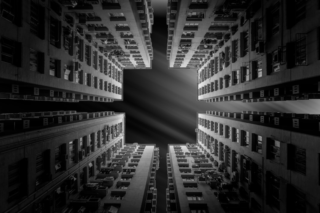 Black and white photography of the symmetrical geometry of buildings in Hong Kong, capturing its architectural beauty from below. The cityscape is seen through the center of two tall apartment blocks, creating an intense contrast between lightness and darkness. A black void forms at both ends of each building’s structure, adding depth to the composition. This scene embodies the cinematic style of Wong Kar Wai with a strong emphasis on urban architecture. –ar 128:85