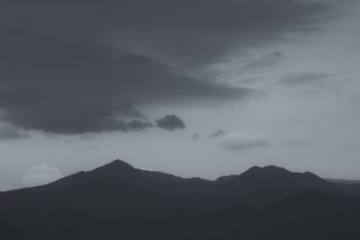 Black and white photograph of mountains with dark clouds, taken from a low angle. The shot has a simple, minimalist and clean aesthetic suitable as a background or wallpaper. The grey sky features a horizon and silhouette of a mountain range and hills in the natural scenery. The tranquil, calm and peaceful scene depicts a tranquil and serene beauty of an evening dusk on a cloudy day with a moody, mysterious and foggy atmosphere, resembling a high resolution and high quality cinematic and realistic photograph in the style of a minimalist nature photographer. --ar 128:85
