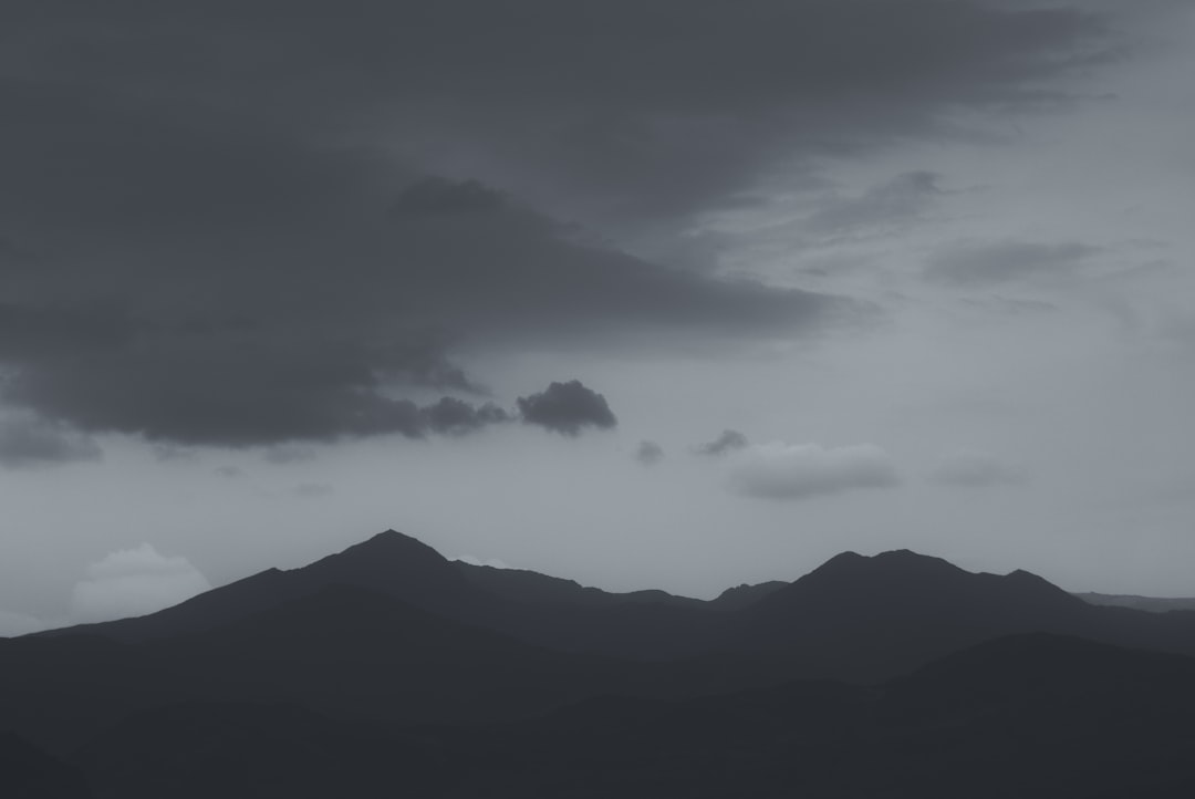 Black and white photograph of mountains with dark clouds, taken from a low angle. The shot has a simple, minimalist and clean aesthetic suitable as a background or wallpaper. The grey sky features a horizon and silhouette of a mountain range and hills in the natural scenery. The tranquil, calm and peaceful scene depicts a tranquil and serene beauty of an evening dusk on a cloudy day with a moody, mysterious and foggy atmosphere, resembling a high resolution and high quality cinematic and realistic photograph in the style of a minimalist nature photographer. –ar 128:85