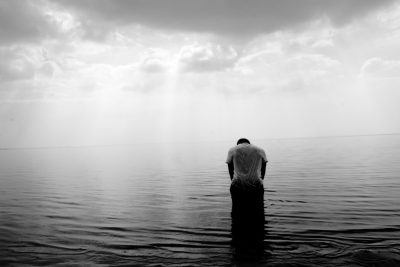 A man is kneeling in the middle of an endless sea, praying to God with his head bowed down and hands clasped together. The sky above him was cloudy, creating a black and white contrast between light gray water ripples and dark clouds. He stood tall against vast calm waters, reflecting subtle colors on their skin, as if he had just been rising from beneath it all along. This scene is depicted in the style of photography with a Leica M6 film camera lens. --ar 128:85