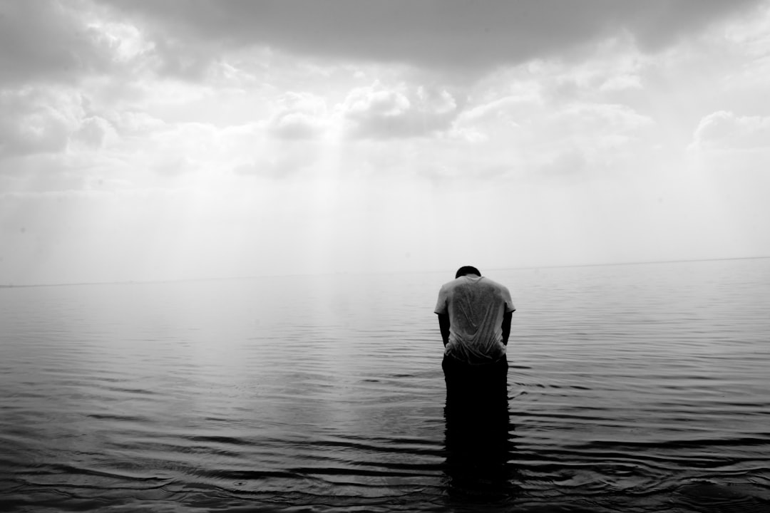 A man is kneeling in the middle of an endless sea, praying to God with his head bowed down and hands clasped together. The sky above him was cloudy, creating a black and white contrast between light gray water ripples and dark clouds. He stood tall against vast calm waters, reflecting subtle colors on their skin, as if he had just been rising from beneath it all along. This scene is depicted in the style of photography with a Leica M6 film camera lens. –ar 128:85