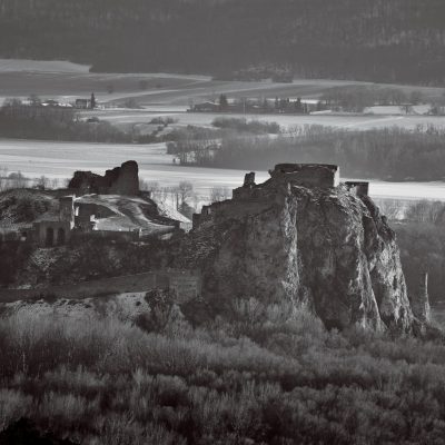 Black and white photograph of the ruins of an ancient castle on top of hill overlooking a valley with medieval villages, late winter, early spring morning, shot from high above