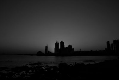 A black and white photograph of the skyline of the city with an ocean in front, silhouette style, at night time, buildings silhouetted against the sky, buildings on the left side only, shot from far away, a beach in the foreground, high contrast, a distant view, a distant cityscape, skyscrapers, a distant shore line, a sea horizon, city lights, an urban landscape, in the style of architectural details, office towers, clear skies, an evening mood. --ar 128:85