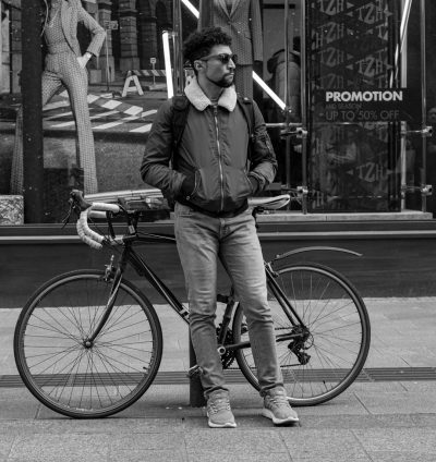 Black and white street photography of an urban young man with his bike in front of the entrance to Zara, wearing jeans, sneakers, glasses and black bomber jacket with fluffy collar. He is standing next to his bicycle leaning against the wall outside the merchant's name - "PFuse" and behind him you can see a large window display featuring models in fashionable outfits, along with signs that say things like 'up to 50% off' or 'promotion'. The atmosphere should feel cool and casual, capturing everyday city life in the style of a street photographer. --ar 15:16