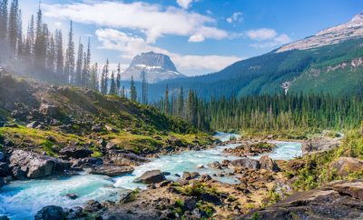 A stunning landscape photograph of the Yosemite National Park in British Columbia, Canada. The scene shows the mountain range with dense forests and rocky peaks, with clear blue skies above. A crystal clear river winds through the valley below, surrounded by lush green trees. In front is an isolated peak that stands tall against the background. Shot on a Nikon D850 DSLR camera using a wide-angle lens to capture the vastness of nature's beauty. The photograph is in the style of [Ansel Adams](https://goo.gl/search?artist%20Ansel%20Adams). --ar 128:77