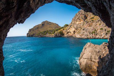 photograph of a mysterious cave on the island of Mallorca with a blue sea and green mountain, view from inside to outside, blue sky, sunny day, in the style of national geographic photography --ar 128:85