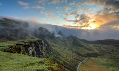 the dramatic highlands of Scotland, neat grassy hills and cliffs with clouds and sun rays breaking through the fog above them, the road leading to an isolated rock outcropping with a sunset in front of it, green, in the style of canon eos r5 --ar 32:19