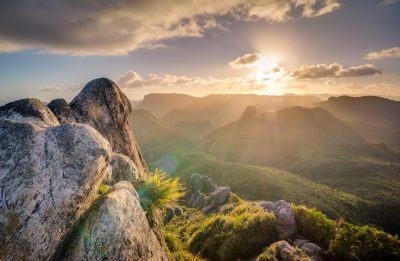 A stunning sunrise over the rugged peaks of Gondangaparagana Mountain, captured from atop its rocky peak in Rio de Janeiro's mountains. Sun rays shining through clouds, golden hour lighting, beautiful green landscape with mountain range in background, highly detailed, professional photography, depth of field, shot on Sony Alpha A7 III camera with Canon EF F/2.8L II USM lens in the style of professional photography. --ar 128:83