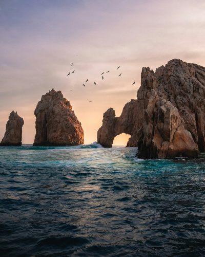 Photo of the arc rock in Los Cabos, Mexico at sunset with birds flying over it. The arch is in focus and there's calm water around it. It should look like an instagram photo --ar 51:64