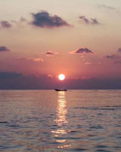 The setting sun shines on the calm sea, reflecting its golden light in the distance. A small boat is floating alone at sunset, with people sitting inside it and enjoying their time in the style of fishing or swimming. The sky above is filled with soft pink clouds that create an atmosphere of tranquility and beauty. Natural scene, photography, telephoto lens. --ar 51:64