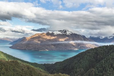 Photograph of Queen's Drift from the top, view over lake and mountains in New Zealand, clouds, blue sky, lake with turquoise water, green forest, snow on mountain tops, shot in the style of Canon EOS R5. --ar 128:85