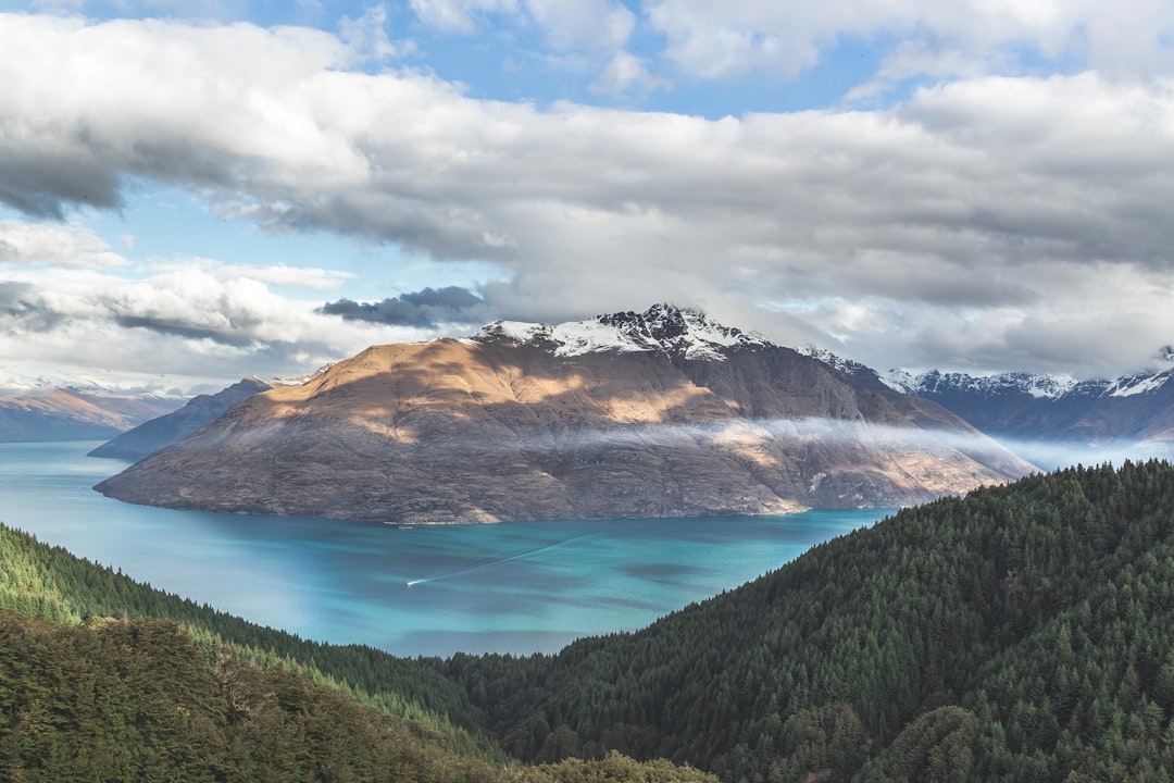 Photograph of Queen’s Drift from the top, view over lake and mountains in New Zealand, clouds, blue sky, lake with turquoise water, green forest, snow on mountain tops, shot in the style of Canon EOS R5. –ar 128:85