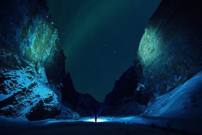 A person standing in the middle of an icy canyon at night, illuminated by the northern lights, with snowcovered cliffs and glowing green moss on both sides. The scene is captured from behind as if taken using Canon eos r5 camera with f/8 aperture setting, creating a dramatic contrast between light and dark colors. A man holding up his flashlight illuminates the ground below him. He's wearing warm  to stay covered against cold weather, and there’s no other people around. --ar 128:85