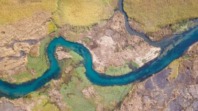 Aerial view of a winding river flowing through marshland, with vibrant blue water between green and brown land, from a top-down perspective, in the style of natural landscape photography, with high resolution and detailed texture. The bird's-eye view shows the natural colors and dynamic flow, in a detailed brush-like style. The overall composition conveys the beauty found in nature's contrast with urban life. --ar 16:9