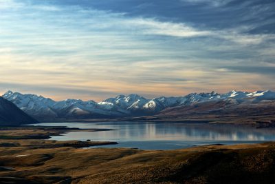 A panoramic view of the mountains and lake at sunset, with snowcapped peaks in New Zealand's outline in the background. The scene is captured from an elevated perspective overlooking Lake glacial water reflecting the sky. A small town sits on one side of the lake, adding to its sense of scale. In the foreground lies flat brown grassland leading up to the lake, creating contrast between natural beauty and human development. Shot in the style of [Ansel Adams](https://goo.gl/search?artist%20Ansel%20Adams) using a Nikon D850 camera and Nikon AFS NIKKOR 24-70mm f/2G ED lens. --ar 128:85