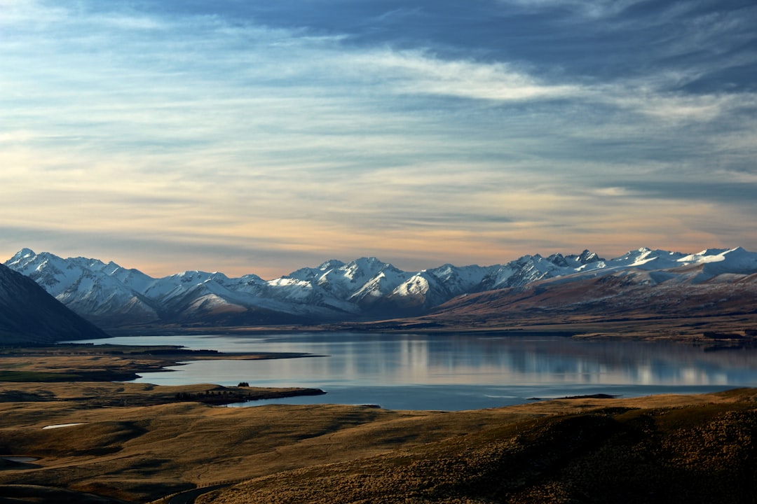 A panoramic view of the mountains and lake at sunset, with snowcapped peaks in New Zealand’s outline in the background. The scene is captured from an elevated perspective overlooking Lake glacial water reflecting the sky. A small town sits on one side of the lake, adding to its sense of scale. In the foreground lies flat brown grassland leading up to the lake, creating contrast between natural beauty and human development. Shot in the style of [Ansel Adams](https://goo.gl/search?artist%20Ansel%20Adams) using a Nikon D850 camera and Nikon AFS NIKKOR 24-70mm f/2G ED lens. –ar 128:85