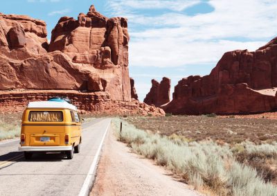 A yellow van driving down the road in Arches National Park, Utah with red rock formations in background. In style of photography, unsplash --ar 64:45