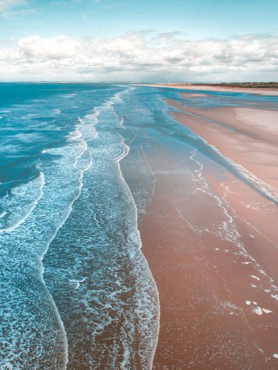 A beautiful beach with waves gently lapping the shore, taken from an aerial perspective. The sand is brown and smooth, while the water has gentle ripples that create long lines of color on its surface. In the distance you can see some land, and above it there is a blue sky with white clouds. This scene gives off a sense of calmness and tranquility, making one feel as if they were standing right next to the sea. --ar 3:4