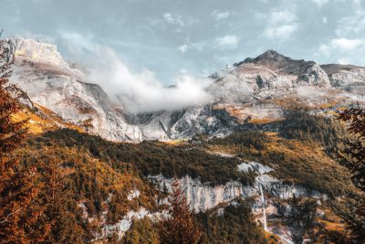 Photograph of the Swiss Alps in autumn, with trees and clouds. The mountain range is visible from below, with white rocks and green vegetation. Shot in the style of Sony Alpha A7R IV at f/8. --ar 128:85