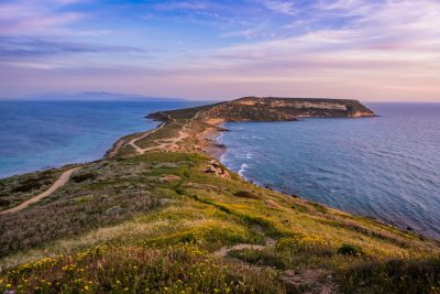 view of the coast from above in Lec فترة, painting with island seen far away in background. The sea is calm and blue, the sky is pastel pink at sunset. Green grass on hillsides, yellow wildflowers along path leading to beach. A small lighthouse can be seen slightly in distance. In the style of National Geographic photo. --ar 128:85