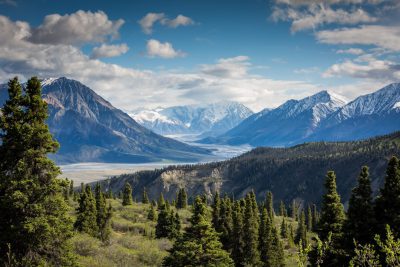 A panoramic view of the Alaska Dynasty, with snowcapped mountains in background and dense forests below. The sky is clear blue with fluffy white clouds, creating an idyllic scene. In front of the camera lies a vast valley filled with lush green trees. A wide river winds through it, adding to its natural beauty. Shot on Canon EOS R5 mirrorless lens --ar 128:85