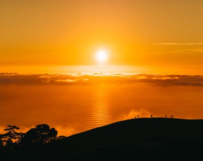 A golden sunset casts an orange glow over the horizon, with silhouettes of people standing on top of hills and trees overlooking the ocean below. The sun is partially obscured by clouds, creating long shadows that add depth to the scene. This tranquil and serene landscape evokes feelings of peace and hope as it reflects tranquility in nature. --ar 128:101