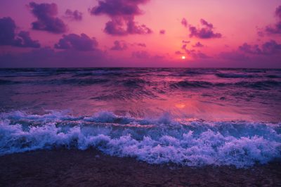 A beautiful purple sunset over the ocean, with waves crashing on shore. The sky is painted in shades of pink and orange as the sun sets behind clouds. A serene scene capturing nature's beauty. High resolution photography, shot in the style of Canon EOS. --ar 128:85