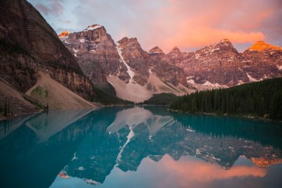 A serene scene of the mountains around Moraine Lake at sunrise, with reflecting blue waters and a pink sky. The surrounding peaks create an epic backdrop for travel photography. The painting depicts the landscape in the style of [Claude Monet](https://goo.gl/search?artist%20Claude%20Monet). --ar 128:85