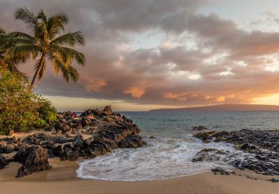 a photo of the beach at sunset in maui, hawaii with palm trees and rocks, waves crashing on shore, people laying down on rock with view over ocean to mountain range in distance, cloudy sky, --ar 128:89