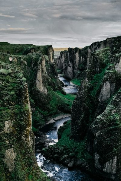 landscape photography of Fjgrown cliffs in Iceland, the green mosscovered rocks tower over the deep chasm below. A river winds through it with a dark sky overhead. The camera captures the majestic view from above, showcasing its rugged beauty and serene atmosphere. In awe, two people stand at one end of the canyon looking down into the waterway. --ar 85:128