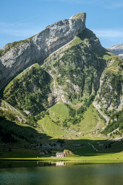 Photo of the Lauterbrunnen mountain in Switzerland, lake and green grass on bottom with small house at foot, blue sky, green mountains, photo taken in the style of Canon EOS R5 f/2 80mm lens --ar 85:128