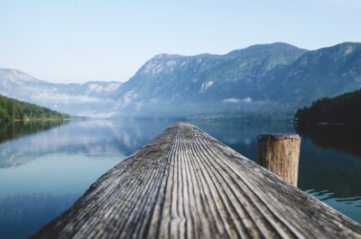 Photograph of the edge of an old wooden dock on Lake Bohinj, looking out at mountains and water. A clear sky on a misty morning. Shot in the style of Sony Alpha A7 III camera for landscape photography. --ar 32:21