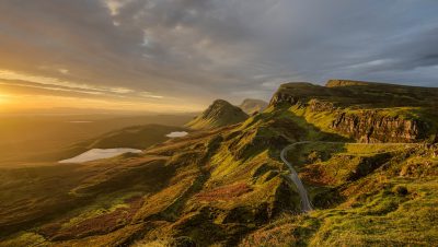 A wide angle view of the Quiraing on the Isle of Skye with a stunning sunrise lighting. The photo was taken from high up on one mountain overlooking another with an S-shaped road winding through it and a lake in the foreground. It is golden hour with warm sunlight casting long shadows over green grassy hills. There are some clouds scattered across the sky and sun rays piercing down onto the water below. The photo is in the style of [Ansel Adams](https://goo.gl/search?artist%20Ansel%20Adams). --ar 16:9