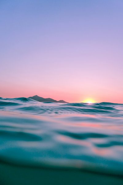 A photograph of the ocean at sunrise, seen from underwater with mountains in the far distance, pink and blue sky, flat horizon line, low angle shot, high definition photography, ultra realistic, in the style of canon eos r5. --ar 85:128