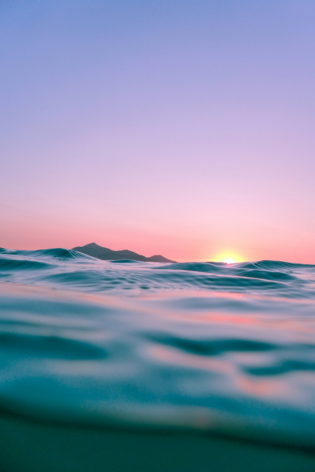 A photograph of the ocean at sunrise, seen from underwater with mountains in the far distance, pink and blue sky, flat horizon line, low angle shot, high definition photography, ultra realistic, in the style of canon eos r5. –ar 85:128