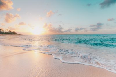 A serene beach scene at sunrise, with soft pastel colors of the sky and turquoise waves gently lapping against the white sand. The sun is just peeking over the horizon casting long shadows on the shore. In the background you can see some tropical trees and in front there's an empty sandy area with gentle surf washing up onto it. This photo was taken using Canon EOS R5 camera. --ar 128:85