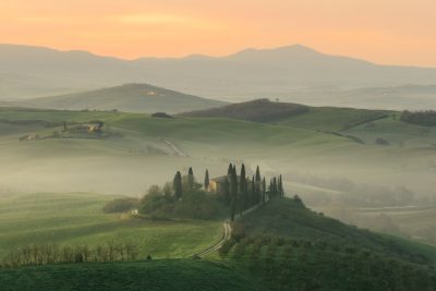 Tuscany, rolling hills and fields at sunrise with mist in the air, soft pastel colors, and an old stone farmhouse surrounded by cypress trees on top of one hill. The valley is filled with green grasses and distant silhouettes of other small buildings. A winding road leads to another house in the distance in the style of an impressionist painter. --ar 128:85