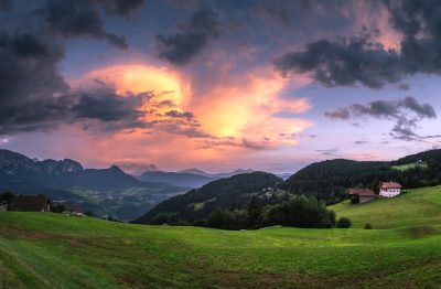 photograph of the Italian Dolomites at sunset, with green grass and dark clouds in the sky, houses on hillsides, mountains visible in background, colorful sky, panoramic view, detailed landscape photography --ar 128:83