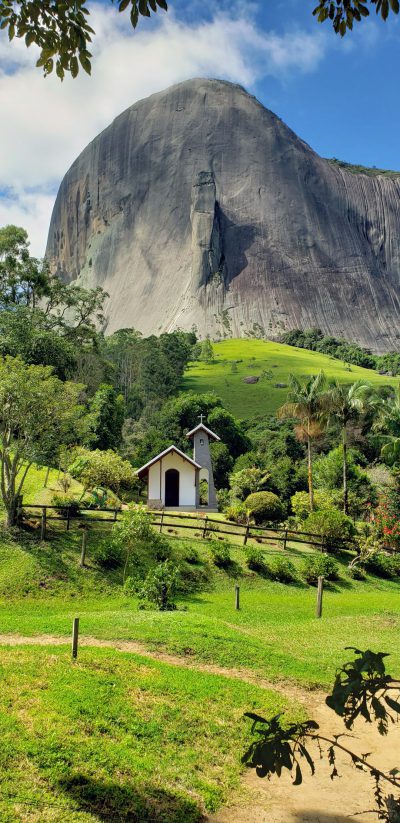 Photo of La Gioconda Photorealistic, high resolution photograph from top to bottom in Brazil with Gu Qi's style rock behind and small church surrounded by green grass near Le Cidir du Mon grand mountain. The photo was taken at an angle with white clouds in the blue sky and trees on both sides. A path leads up towards Gu Wei's style Rock. In front is open space with lawn, surrounded by fence walls. On one side you can see tiny huts. This place seems like paradise. --ar 31:64