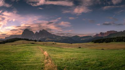 A wideangle view of the Dolomites in Italy at sunset, with grassy fields and mountains under a sky painted in the style of light clouds. The foreground features an old stone path leading to distant peaks, while small structures can be seen scattered across the landscape below. This scene captures nature's beauty and tranquility, offering panoramic views of vast green meadows, towering mountain spires, and serene twilight hues. --ar 16:9