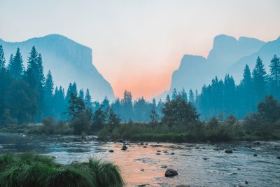 A beautiful landscape photograph of Yosemite National Park at sunrise, with El Capitan and Half Dome in view, taken from a river bed with mist rising off its surface. The sky is a soft pastel pink and blue gradient with some cloud cover. In front there is lush green grass and trees, and behind them towering mountains covered in dense forest. There are rocks visible on both sides of the river, and a small rocky island or outcrop right in the middle of it. --ar 128:85